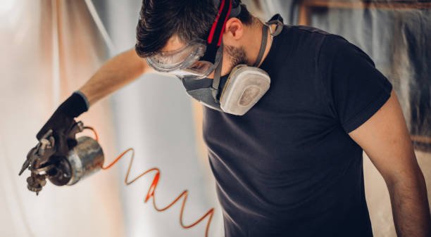 صورة لرجل بقميص أسود يرتدي قفازات ونظارات واقية ومقوى الوجه، يقوم باستخدام بندقية رش موصولة بأنبوب برتقالي داخل غرفة، مما يوضح اتباعه لإجراءات السلامة أثناء العمل.